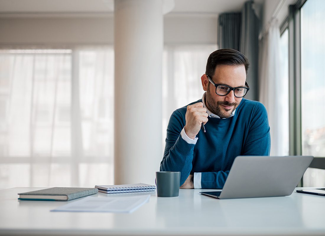 About Our Agency - Business Professional Sitting at His Desk Reading on His Laptop While Holding a Pen